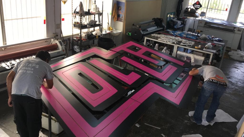 Dos hombres en un taller gráfico montando un gran letrero geométrico negro y rosa.