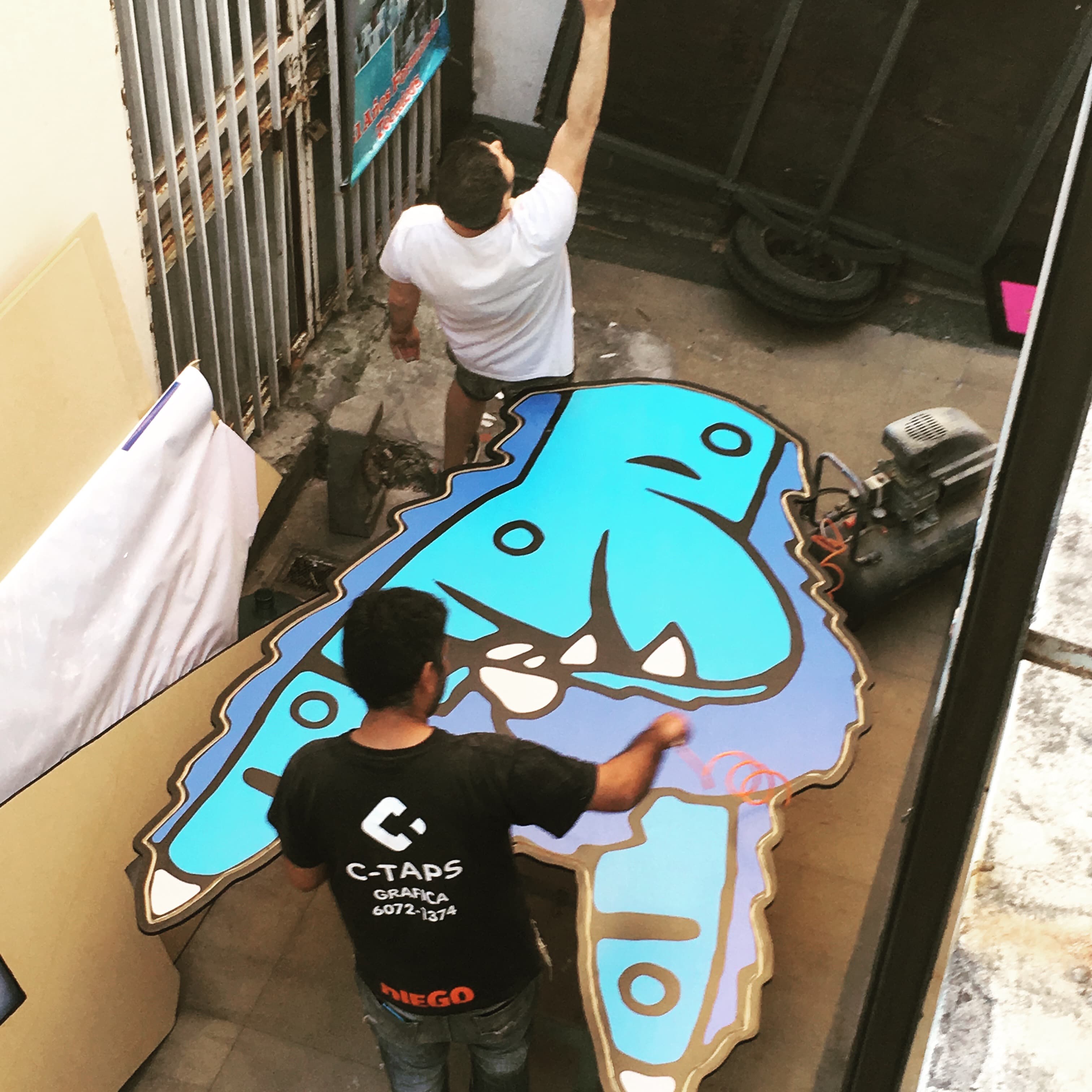 Dos hombres pintando una gran figura azul de un personaje animado en un taller.