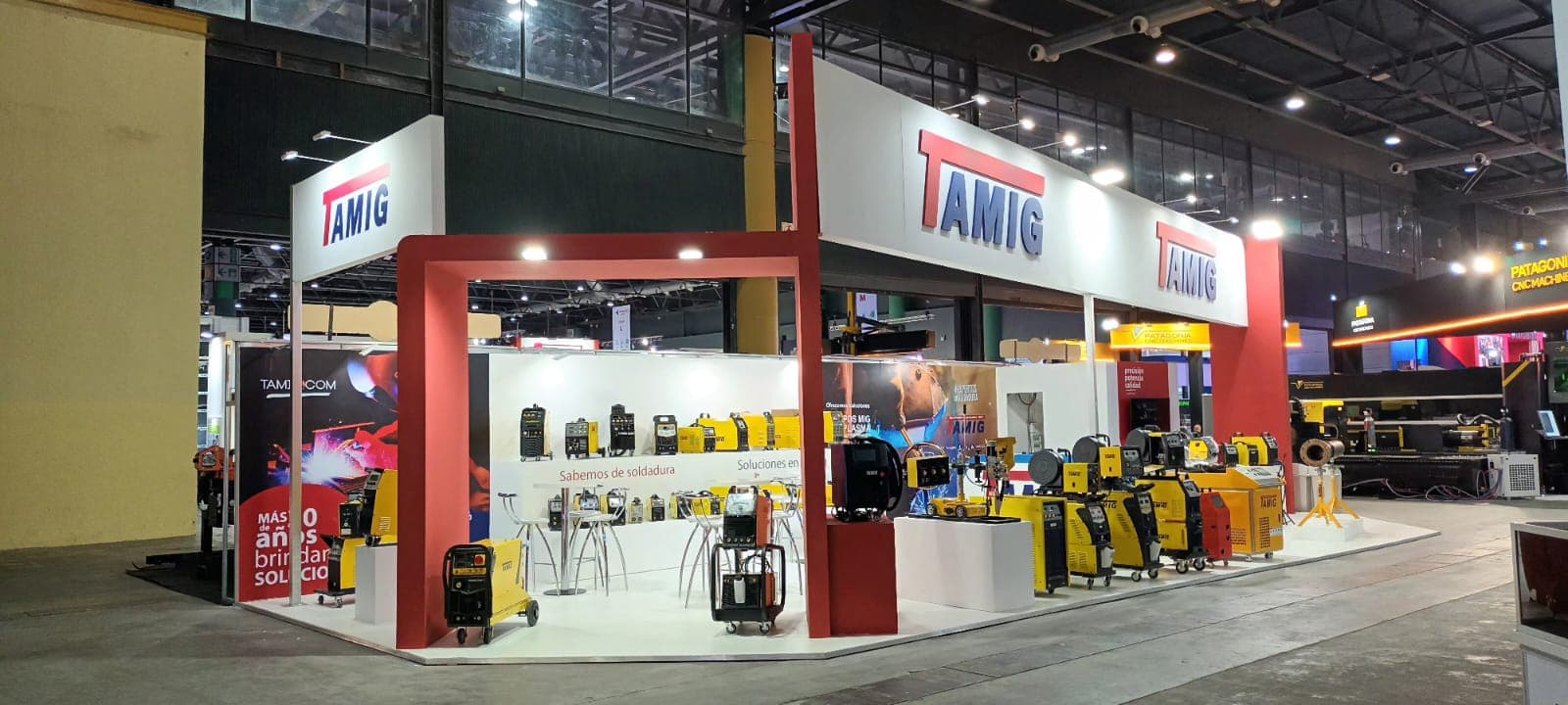 Stand de TAMIG en una feria industrial exhibiendo equipos de soldadura amarillos y negros.