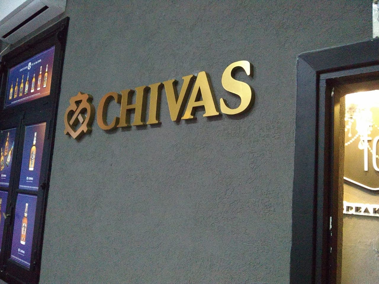 Letras doradas de Chivas y logotipo en relieve sobre una pared gris con carteles publicitarios.