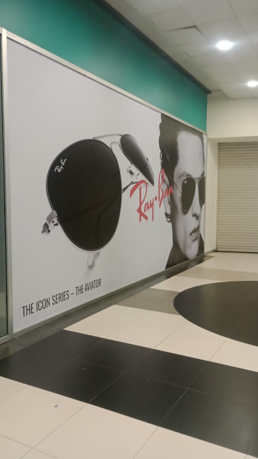 Publicidad de Ray-Ban con gafas Aviator y modelo en un pasillo con suelo ajedrezado.