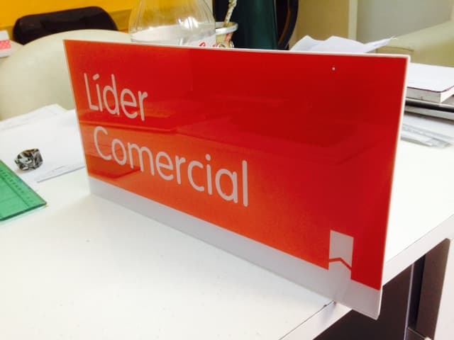 Letrero rectangular rojo con texto blanco "Líder Comercial" sobre una mesa de oficina.